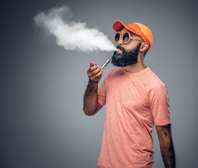 Bearded man in sunglasses and an orange cap exhaling vapor while using a vape pen.
