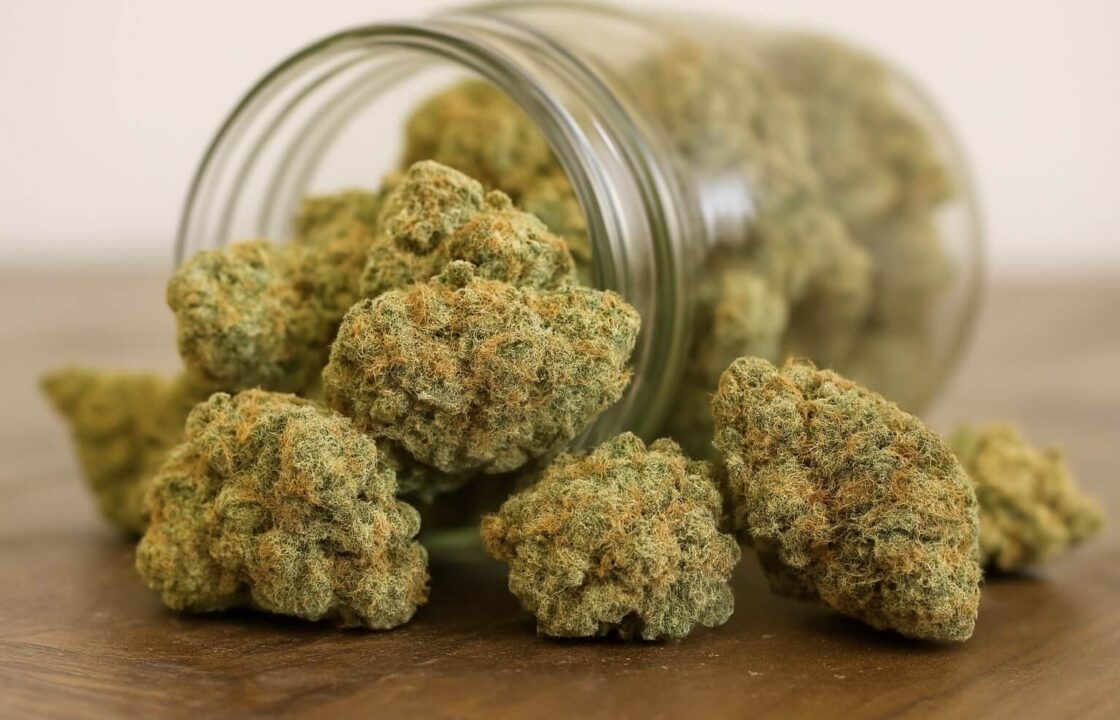 A close-up of cannabis buds spilling from a glass jar onto a wooden surface.