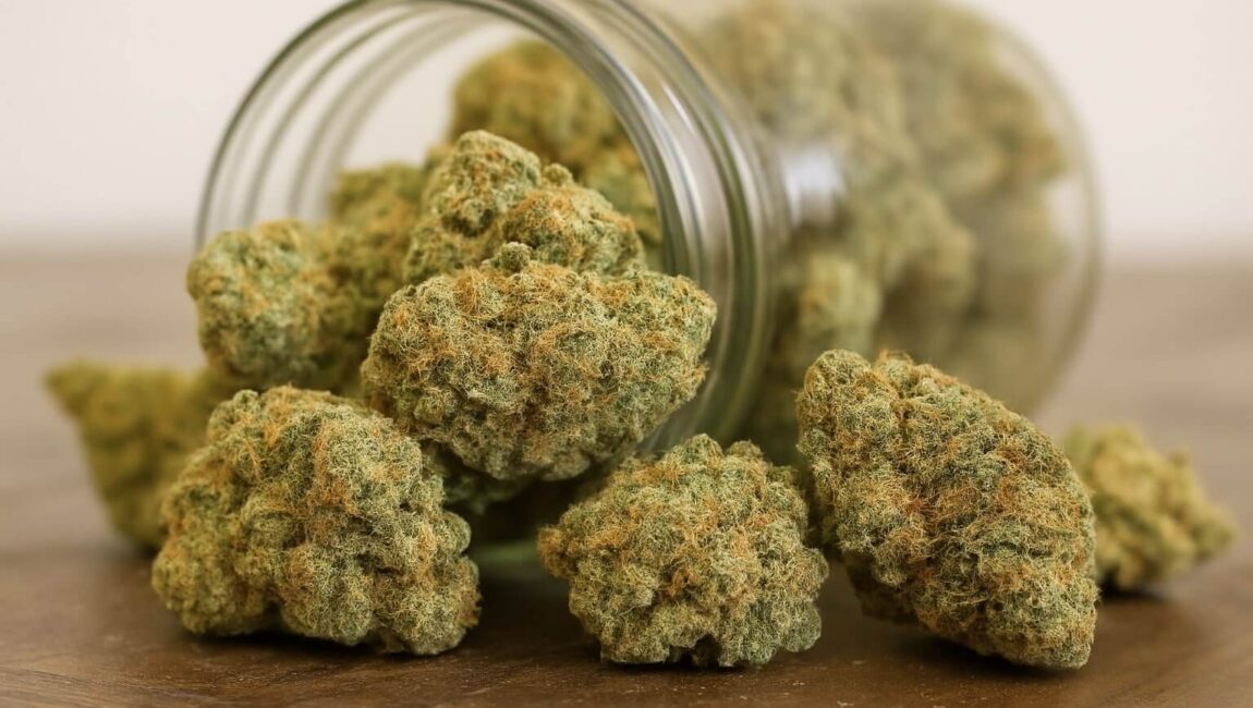 A close-up of cannabis buds spilling from a glass jar onto a wooden surface.