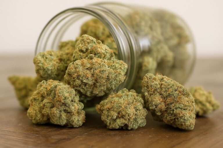 A close-up of cannabis buds spilling from a glass jar onto a wooden surface.
